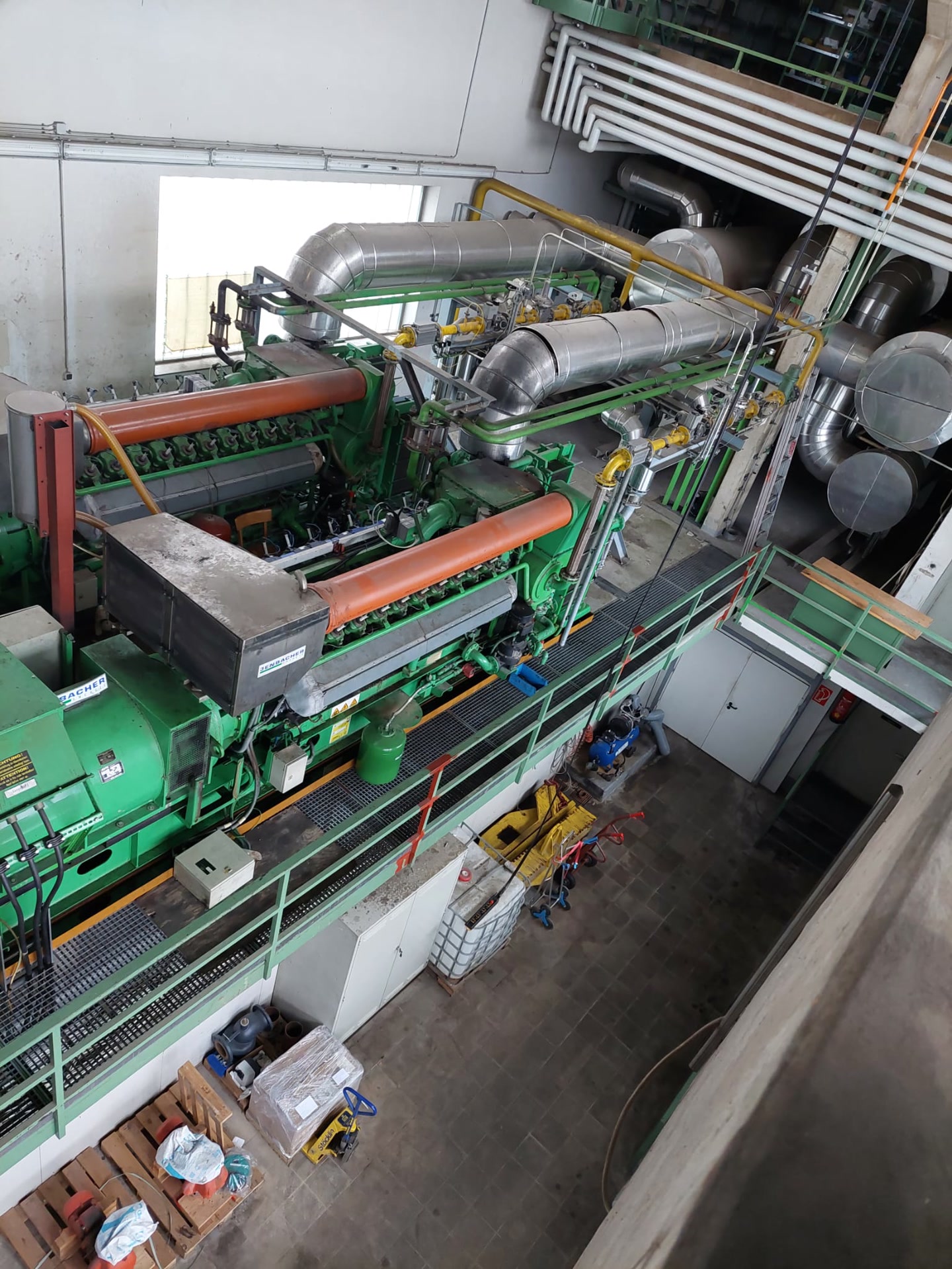 Großer Gasmotor mit Generator auf einem Hallenpodest, Abgasrohre noch angeschlossen.